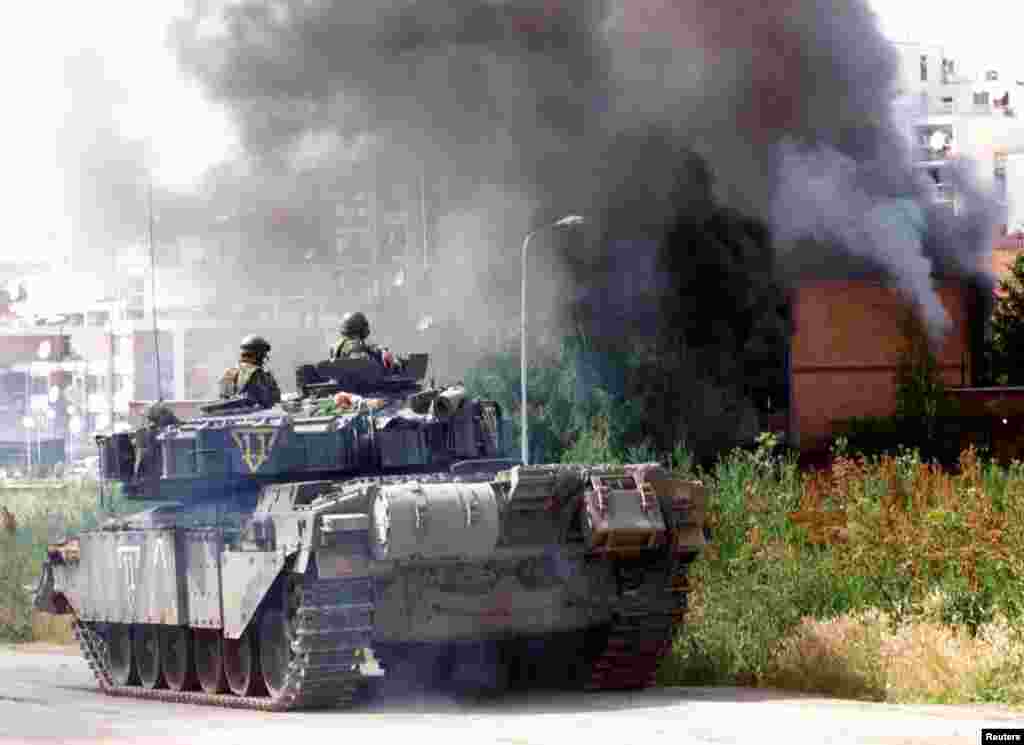 Britanski vojnici gledaju kako kuće gore u Prištini 13. juna 1999. godine, nakon što je, prema Reutersu, na njih pucao srpski vojnik. Nova škola i brojni objekti danas se nižu uz cestu na kojoj su nekada gorjele kuće.